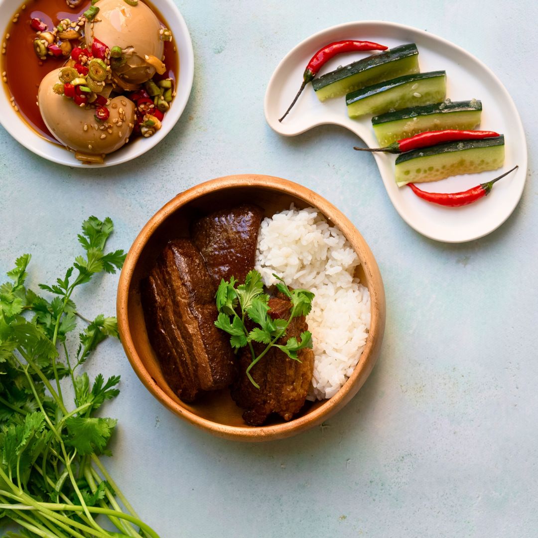 Bowl of food with rice and meat, accompanied by a plate of pickled cucumbers and red peppers on a light blue background.