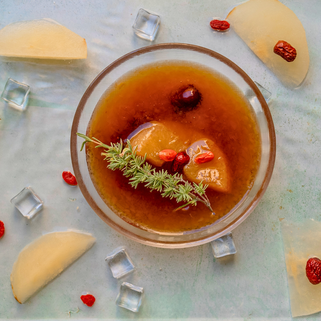 Crystal Pear soup with a brown liquid, slices of fruit, and a sprig of mint on a light blue background.