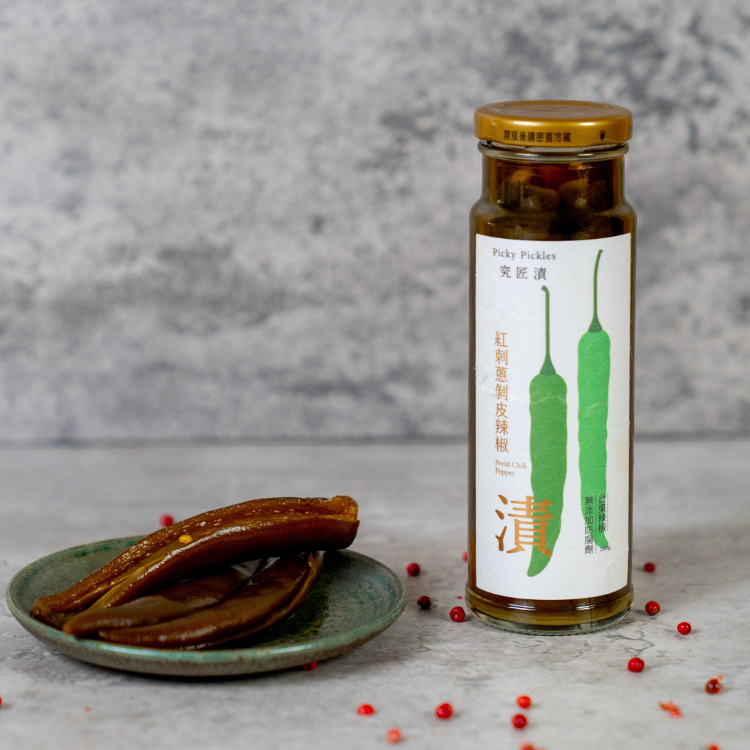 Picky Pickles Peeled Chili Peppers displayed on a green ceramic plate next to the product jar, surrounded by scattered pink peppercorns on a stone tabletop.