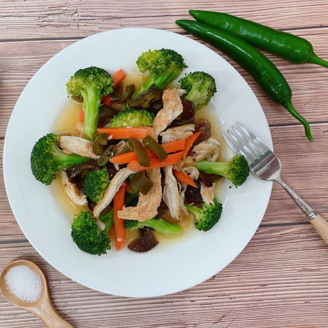 Stir-fried broccoli, shredded chicken, carrots, and mushrooms garnished with chopped peeled chili peppers, plated on a white dish next to whole green chilies and a wooden spoon of salt.