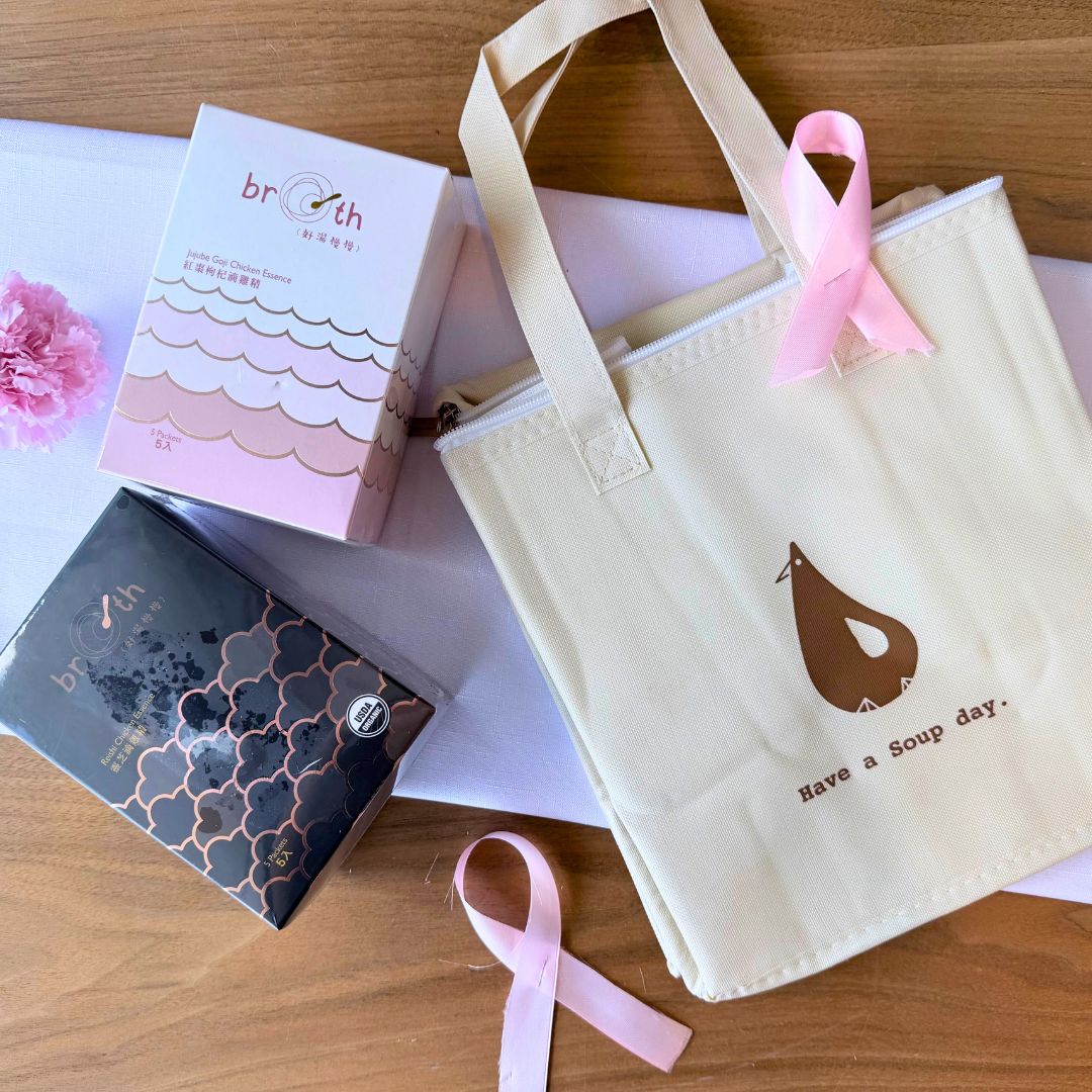 A close-up of the cream-colored BROTH insulated tote bag adorned with a pink ribbon and surrounded by soft pink and white flowers, styled to highlight the warmth and care of the Pink October campaign.