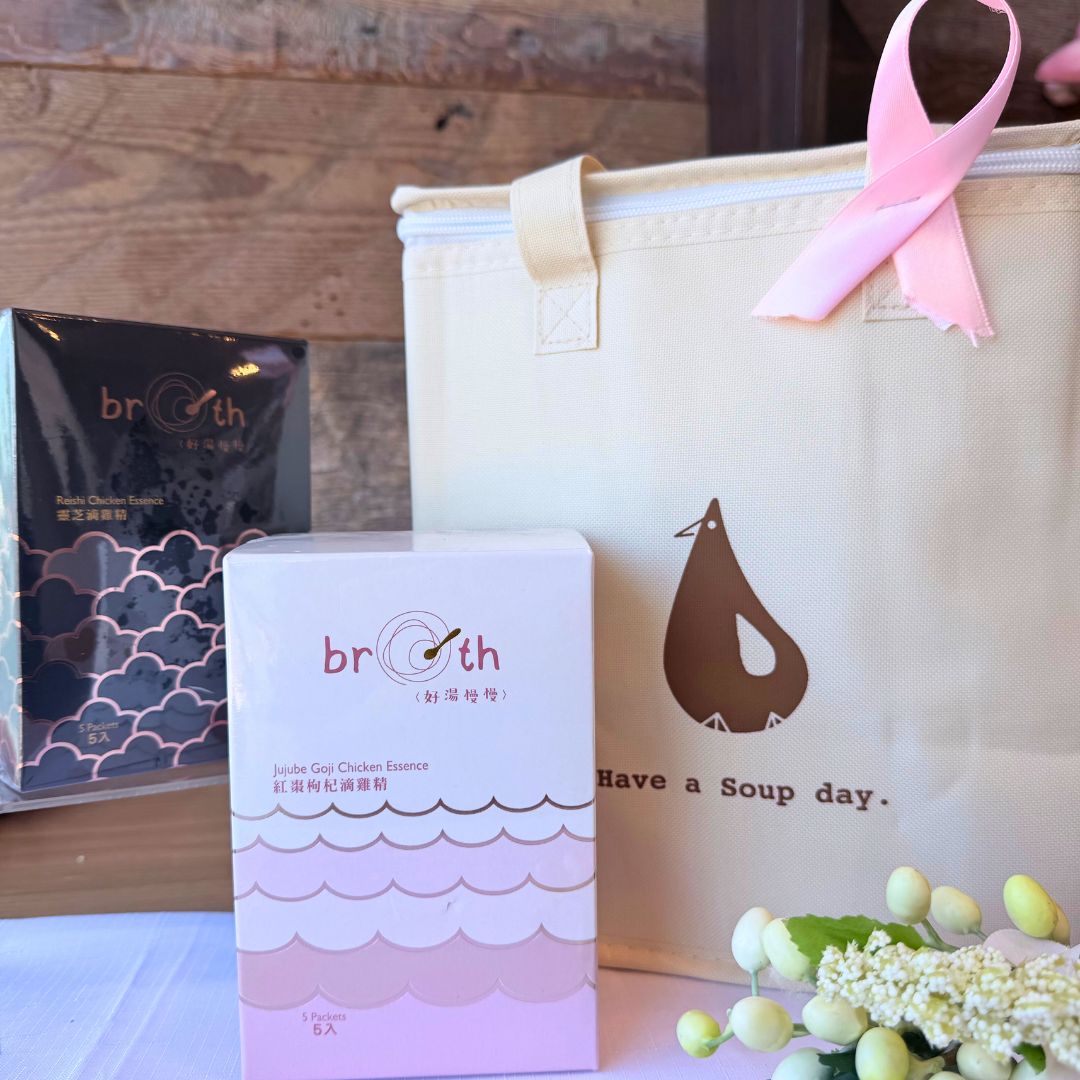 A cozy tabletop scene showing the BROTH insulated tote bag with a pink ribbon and two Chicken Essence gift boxes (Goji Jujube & Reishi) in front. The wooden background adds a rustic, comforting vibe to the Pink October wellness message.
