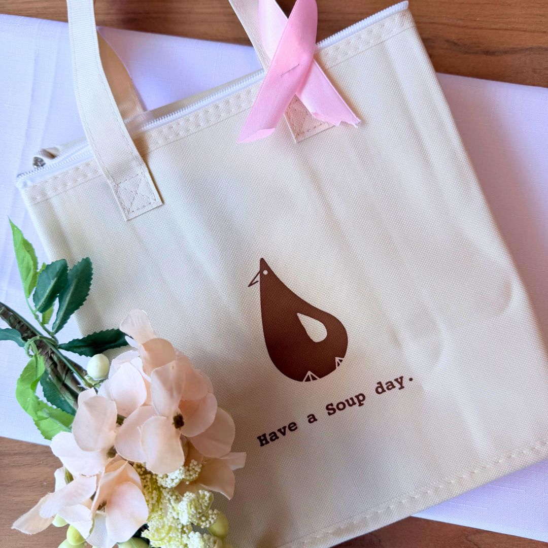 A flat-lay of BROTH’s bundle featuring a cream-colored insulated tote bag with “Have a Soup day” printed on it, paired with two boxes of Chicken Essence—one in pink (Jujube Goji) and one in black (Reishi). A pink ribbon is attached to the tote, symbolizing breast cancer awareness.