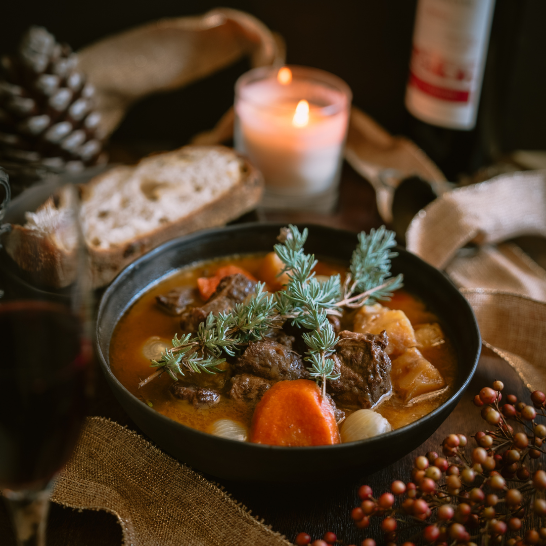 A warm bowl of Beef Bourguignon filled with tender beef, carrots, potatoes, and pearl onions, garnished with fresh herbs, set on a rustic table with a lit candle, bread slices, pinecones, and festive ribbon for a cozy holiday atmosphere.
