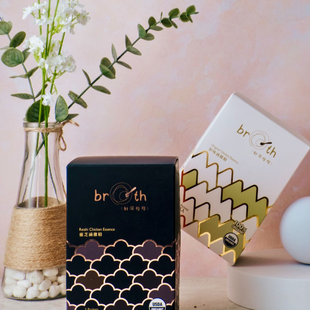 Two boxes of BROTH organic chicken essence(Black: Reishi; Green: orginal) with a vase of flowers on a light background