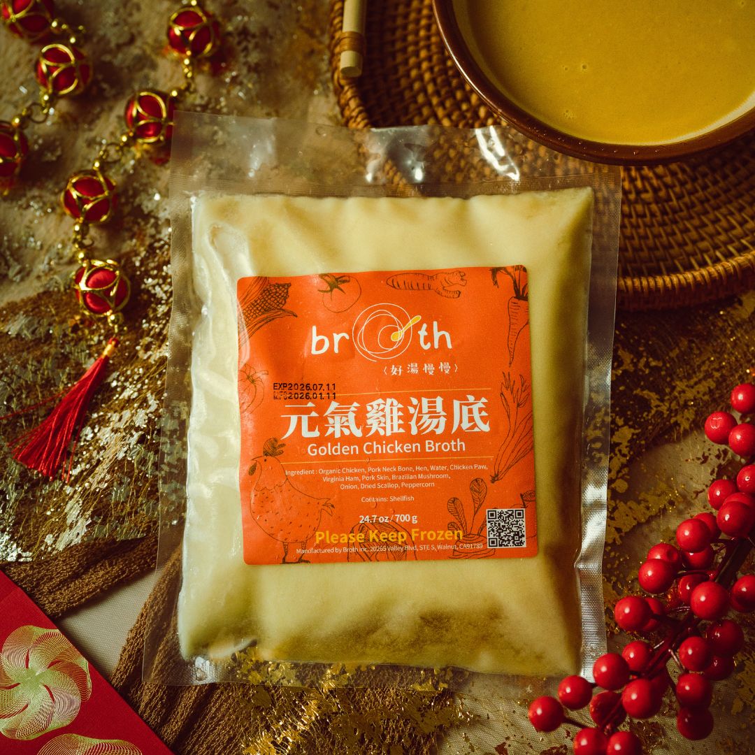 A frozen pack of Golden Chicken Broth from Broth (好湯慢慢) with an orange label, styled on a festive gold background with red tassels, berries, and a bowl of broth to highlight a warm holiday presentation.