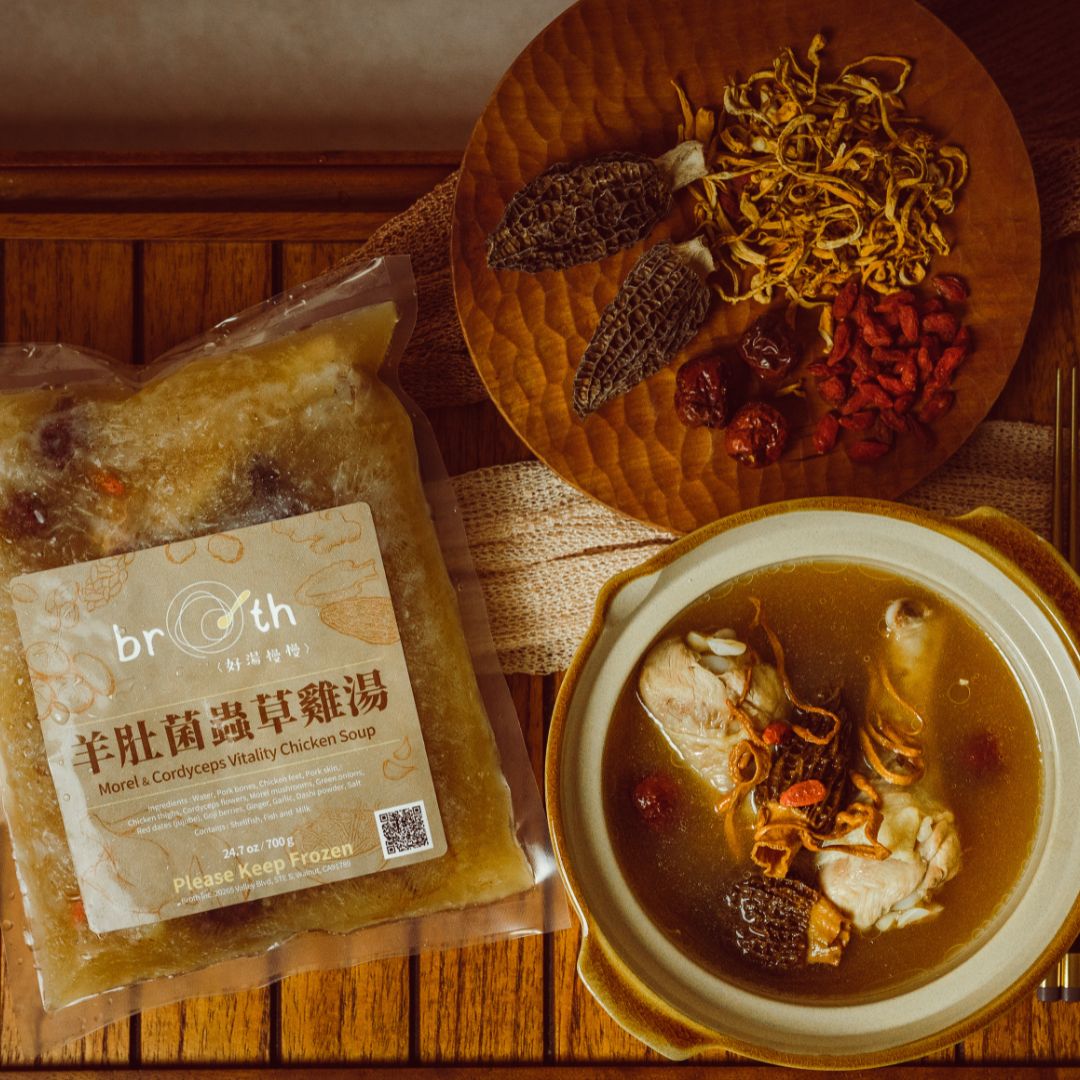 A frozen pack of Morel & Cordyceps Vitality Chicken Soup displayed beside a prepared bowl and dried ingredients, styled on a wooden table.