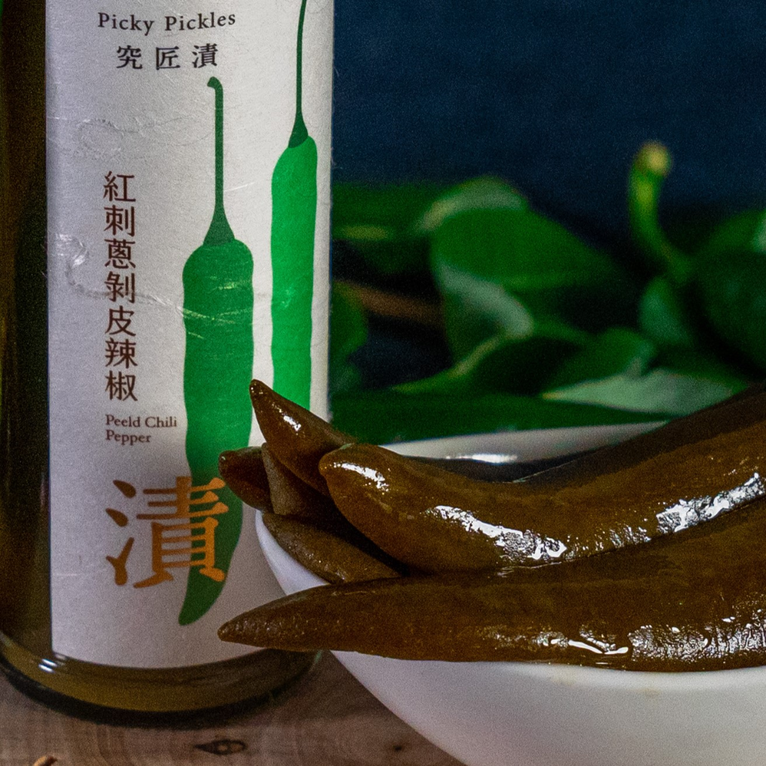 Close-up of Picky Pickles' Peeled Chili Peppers served in a white bowl, with the product jar in the background featuring minimalist green chili illustrations and Chinese calligraphy.