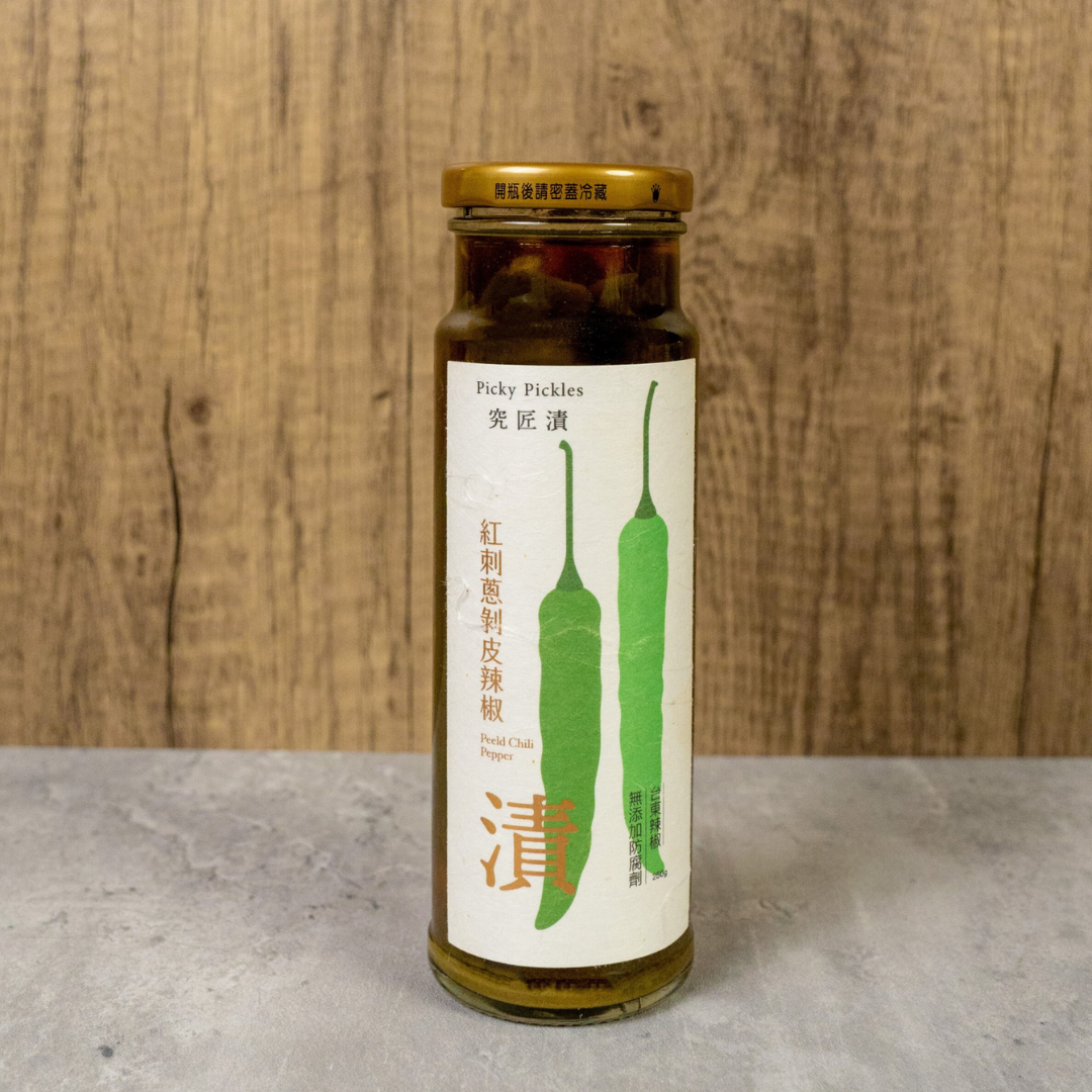 A full product shot of Picky Pickles' Peeled Chili Pepper in a tall glass jar with a golden lid, set against a wooden background.