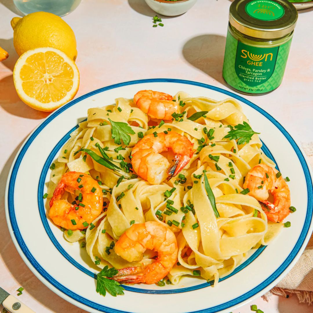 A plate of fettuccine pasta topped with shrimp and garnished with fresh herbs, paired with Sun Ghee Chives, Parsley & Tarragon clarified butter in the background.