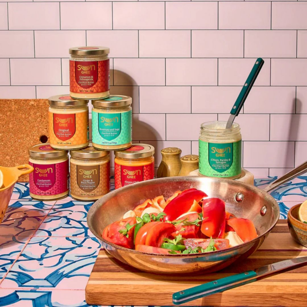 A colorful kitchen scene with a jar of Sun Ghee’s “Chive, Parsley & Garlic” flavor open with a spoon, surrounded by a variety of Sun Ghee jars in different flavors. A sauté pan with tomatoes and herbs is being cooked on the stove, showcasing ghee in culinary use.