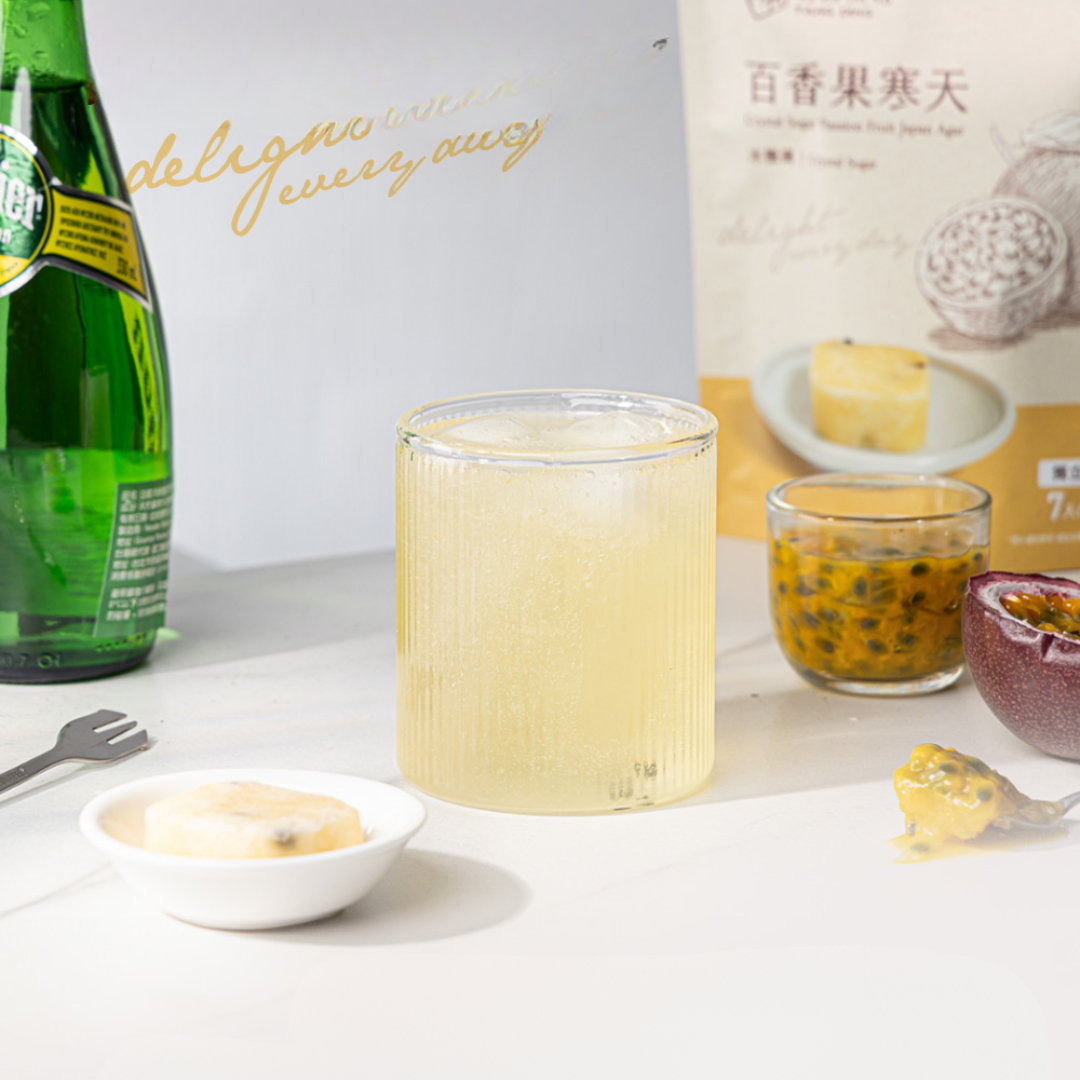 Bottle of sprinkling water, glass of lemonade, and passion fruit on a white surface with a box in the background.