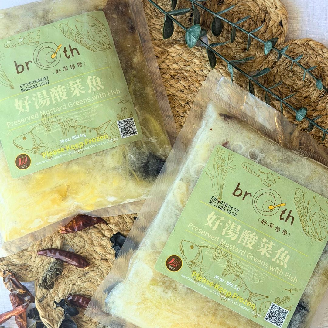 Two packs of BROTH’s Preserved Mustard Greens with Fish placed on a woven mat with dried chilies and herbs, showcasing the product’s natural texture in a warm, cozy setting.