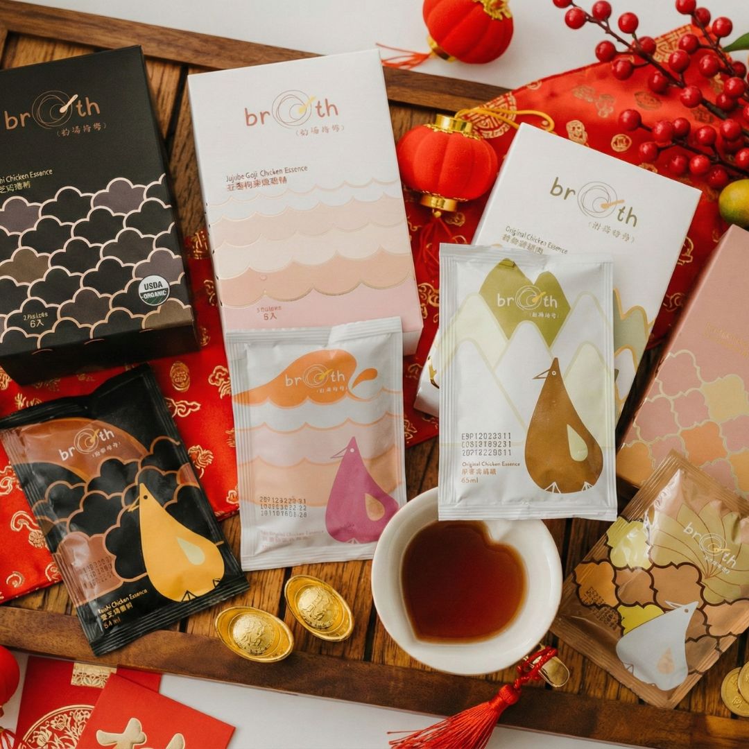 A flat-lay of Broth (好湯慢慢) chicken essence gift sets and sachets alongside a small bowl of chicken essence, red envelopes, and festive Chinese New Year elements.