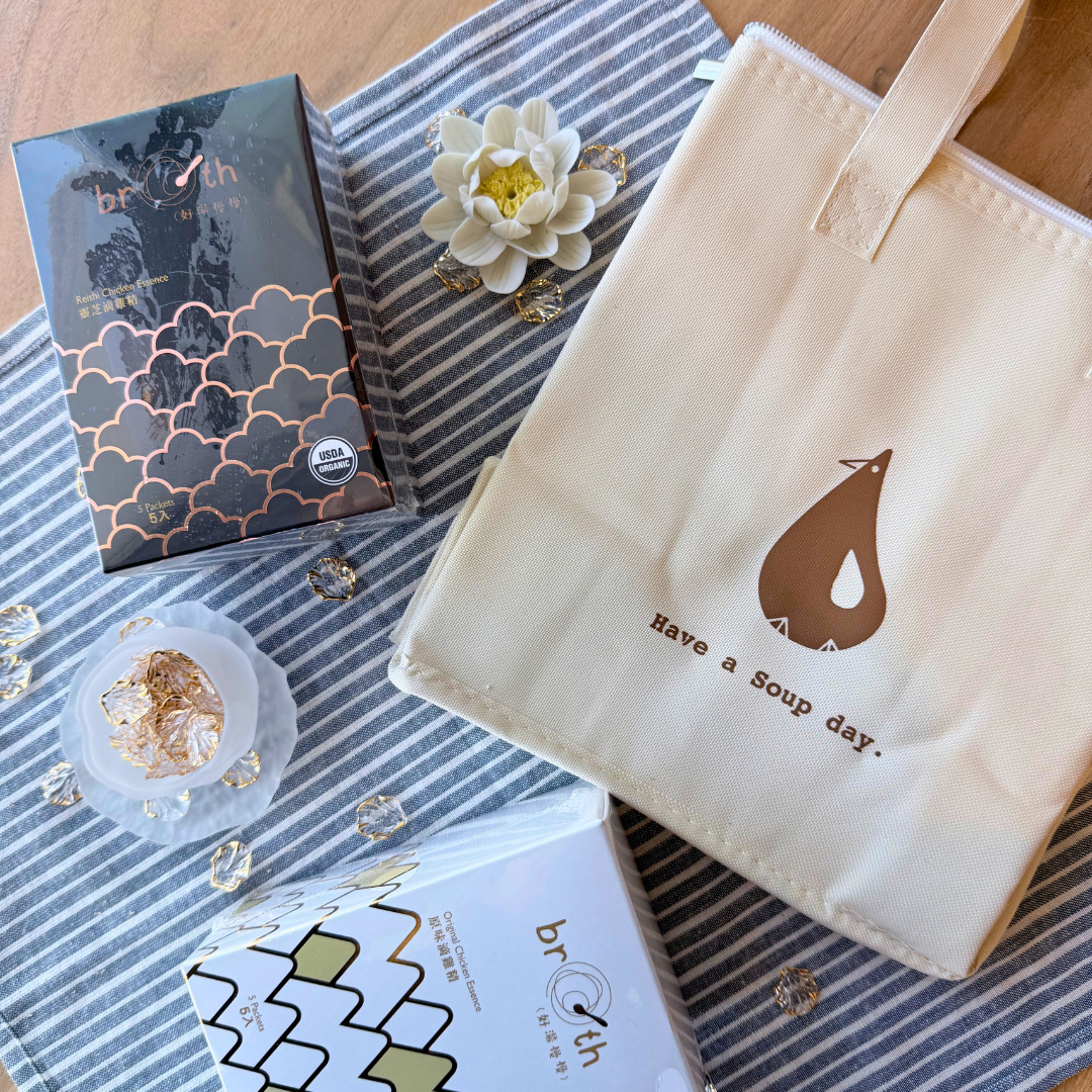 Flat-lay image of Reishi Chicken Essence and Original Chicken Essence boxes beside a striped fabric and a cream cooler bag with the “Have a Soup day” logo. Decorative crystal beads and a lotus flower add a festive touch.
