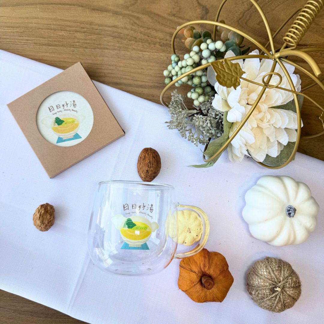 Flat-lay of BROTH merch set featuring a double wall glass mug and diatomite coaster with soup bowl illustration, surrounded by nuts, flowers, and autumn décor.
