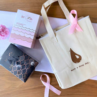 A close-up of the cream-colored BROTH insulated tote bag adorned with a pink ribbon and surrounded by soft pink and white flowers, styled to highlight the warmth and care of the Pink October campaign.