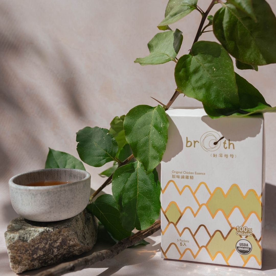 A white and yellow patterned box of breath herbal tea stands next to a small ceramic cup on a stone surface, with green leaves and branches draped over the scene in a styled studio setup.