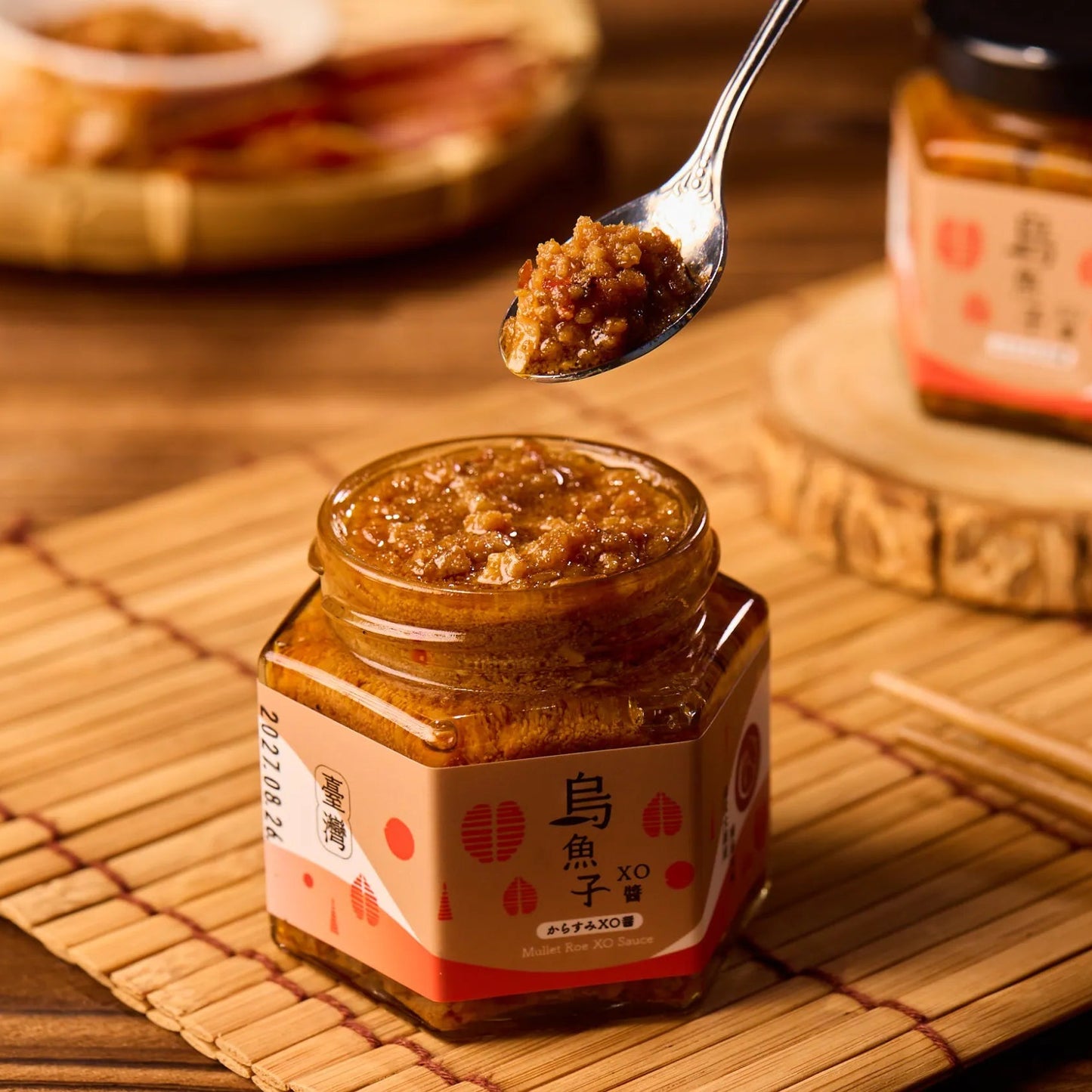 A spoon lifting a scoop of 【The Carp】Mullet Roe XO Sauce from an open jar, showcasing the sauce’s chunky texture and glossy finish on a bamboo mat.